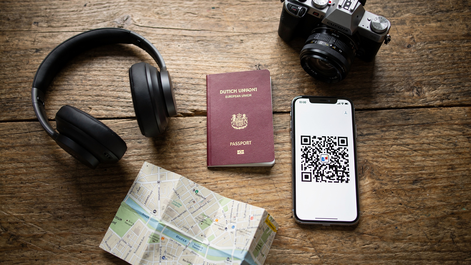 Een overzichtsfoto van een travel-flatlay met een open paspoort, een smartphone met een QR-code op het scherm, een paar koptelefoons, een compacte camera en een reiskaart, allemaal netjes gerangschikt op een houten tafel, klaar voor vertrek.