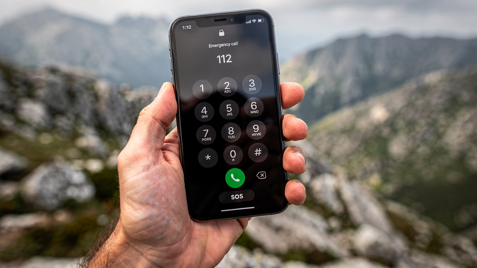 Uma mão segura um smartphone com uma interface de chamada de emergência no ecrã, com um fundo desfocado de uma paisagem montanhosa, sugerindo a importância de estar conectado em áreas remotas.