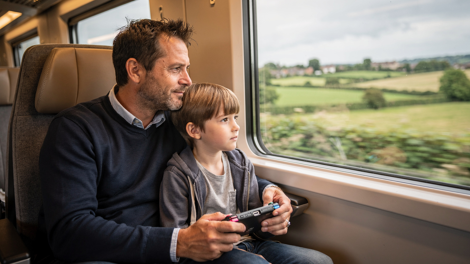 Un père et son fils regardant par la fenêtre d'un train traversant la campagne anglaise, le fils tenant une console de jeu portable.