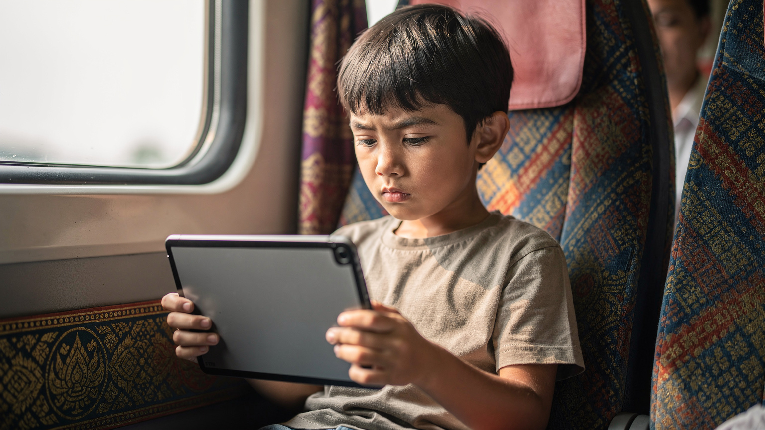 A young child, around 7 years old, looking dejectedly at a blank tablet screen while seated on a vibrant, modern train in Thailand, indicating a loss of connectivity or drained battery, with a slightly worried parent in the soft-focus background.