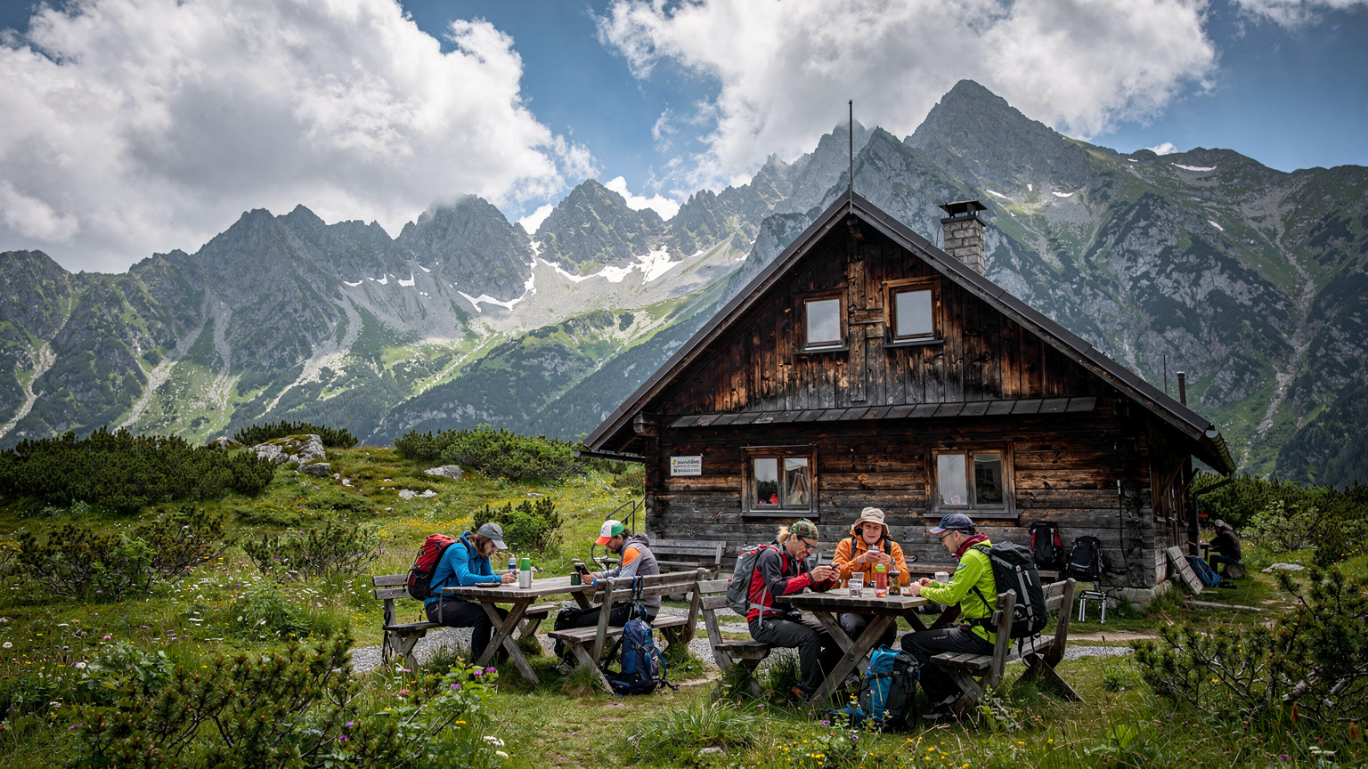 Záber na horskú chatu v slovenských Tatrách, obklopenú zelenými lúkami a skalnatými štítmi. Pred chatou sedia turisti a sledujú svoje telefóny, pravdepodobne kontrolujú signál.