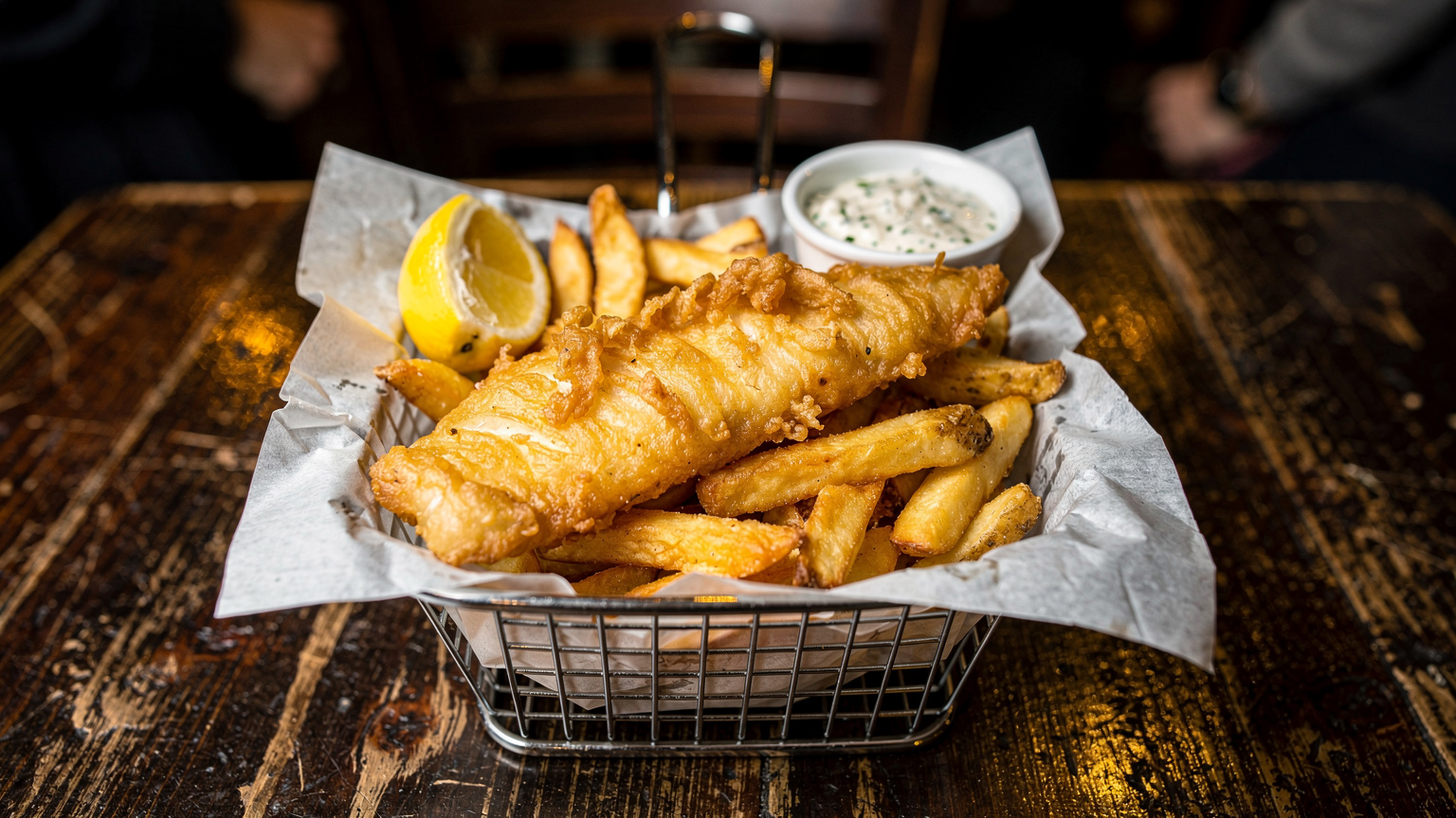 Классическое британское блюдо Fish and Chips, поданное в корзинке с соусом тартар, на деревянном столе в лондонском пабе.
