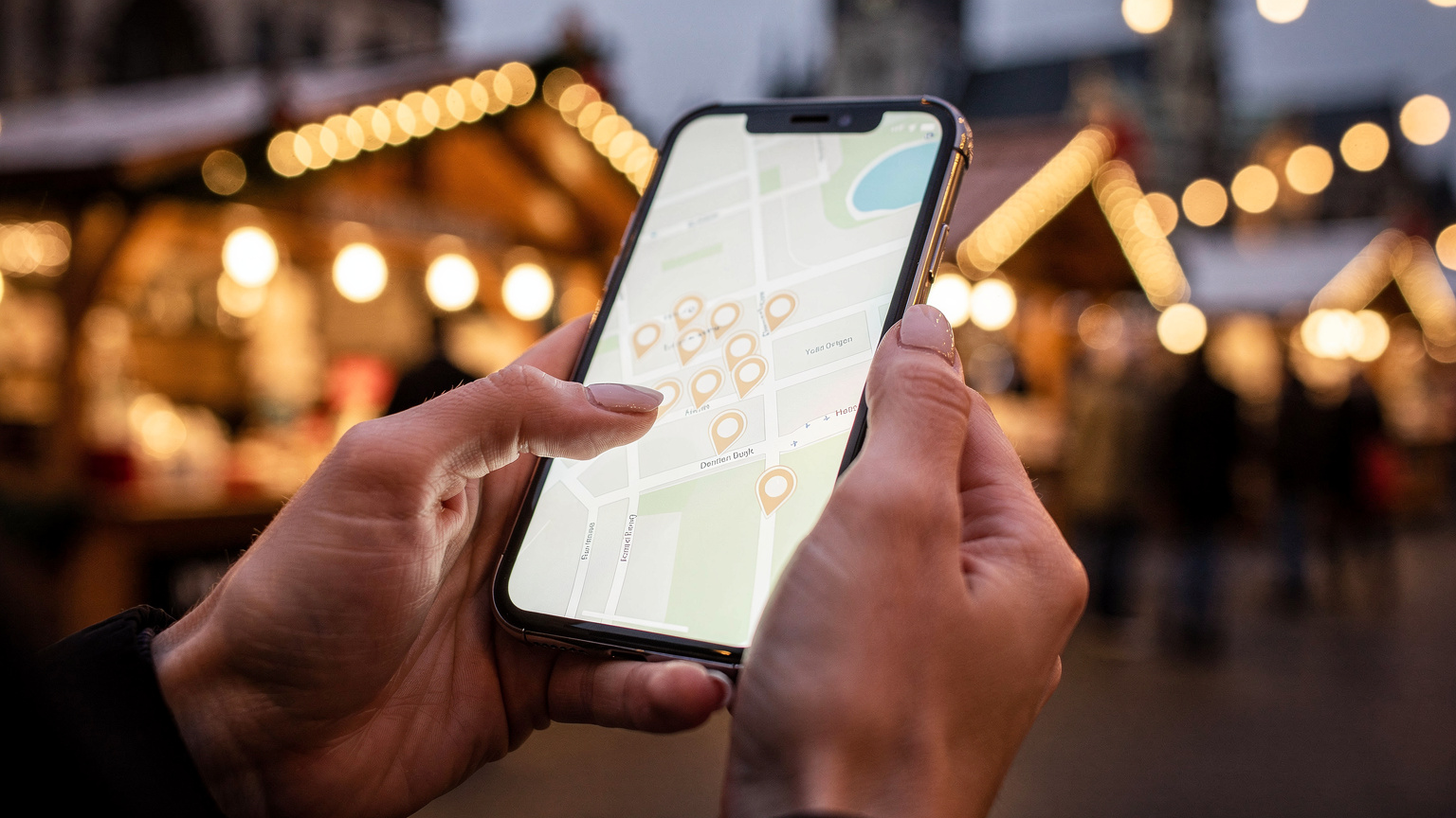 Eine Nahaufnahme zeigt Hände, die ein Smartphone halten, auf dem eine Karte des Weihnachtsmarktes in Köln angezeigt wird, im Hintergrund sind verschwommen die festlichen Lichter zu sehen.