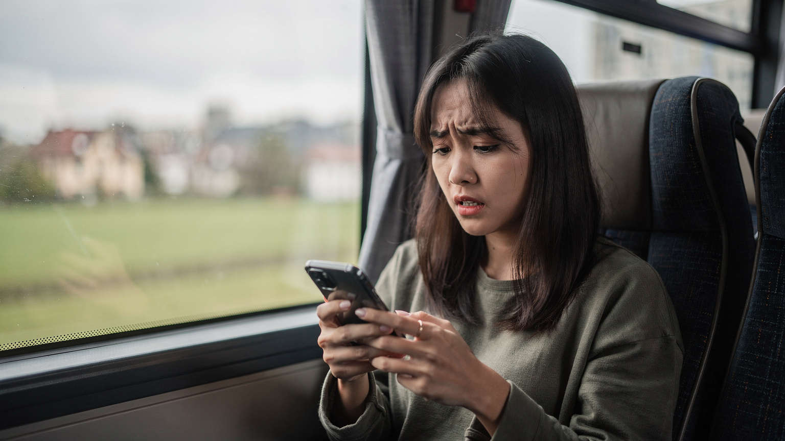 Một phụ nữ trẻ đang tỏ vẻ lo lắng, nhìn chằm chằm vào màn hình điện thoại của mình trong khi đang ngồi trên một chiếc xe buýt ở Châu Âu, có lẽ đang gặp vấn đề về kết nối mạng.