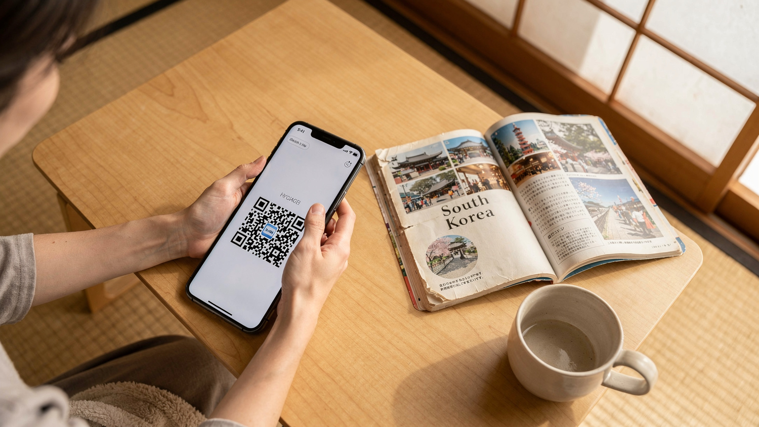 日本の自宅のリビングで、母親がスマートフォンをテーブルに置き、別のスマートフォンでeSIM設定のQRコードをスキャンしている様子。そばには開かれた韓国の旅行ガイドブックが置かれている。