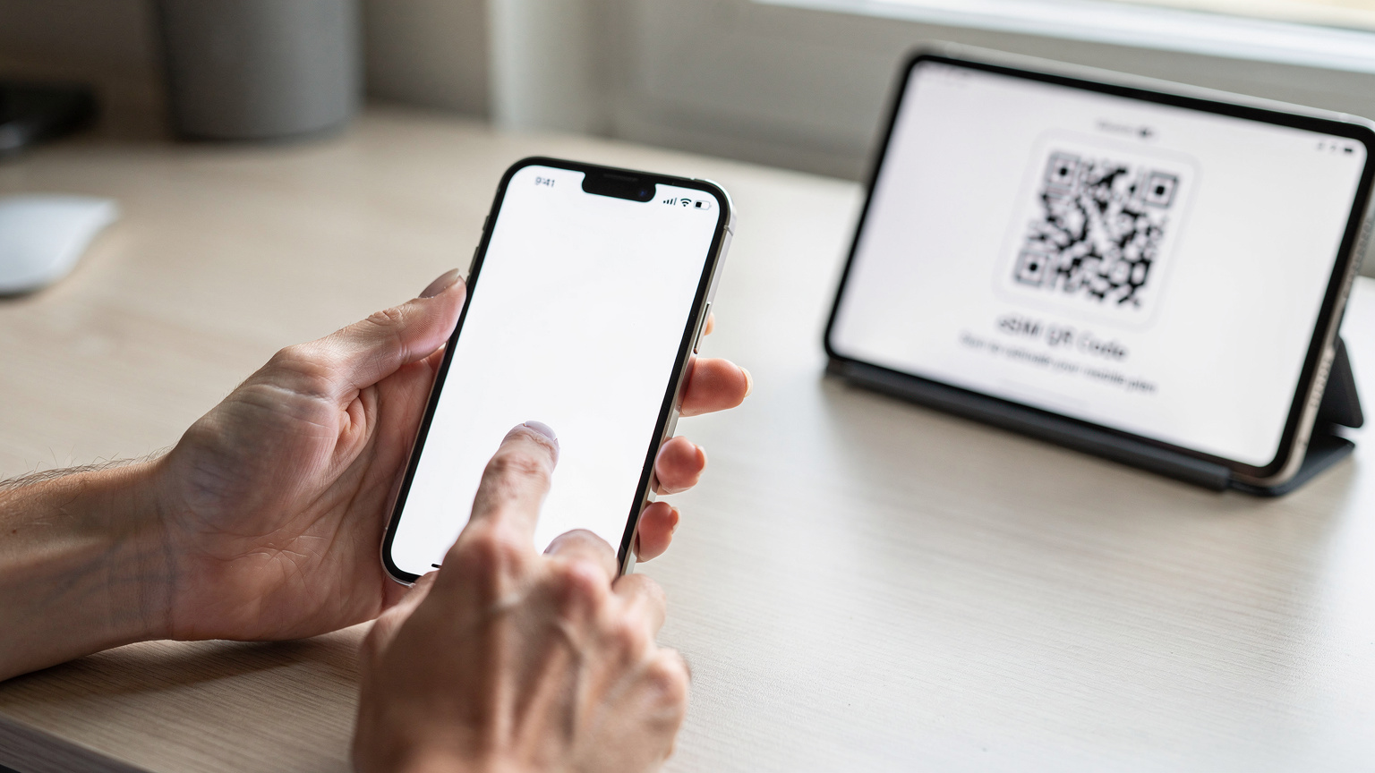 Mani eleganti che regolano le impostazioni della rete cellulare su uno smartphone iPhone 15, con una guida utente aperta su un tablet sullo sfondo che mostra un codice QR eSIM.
