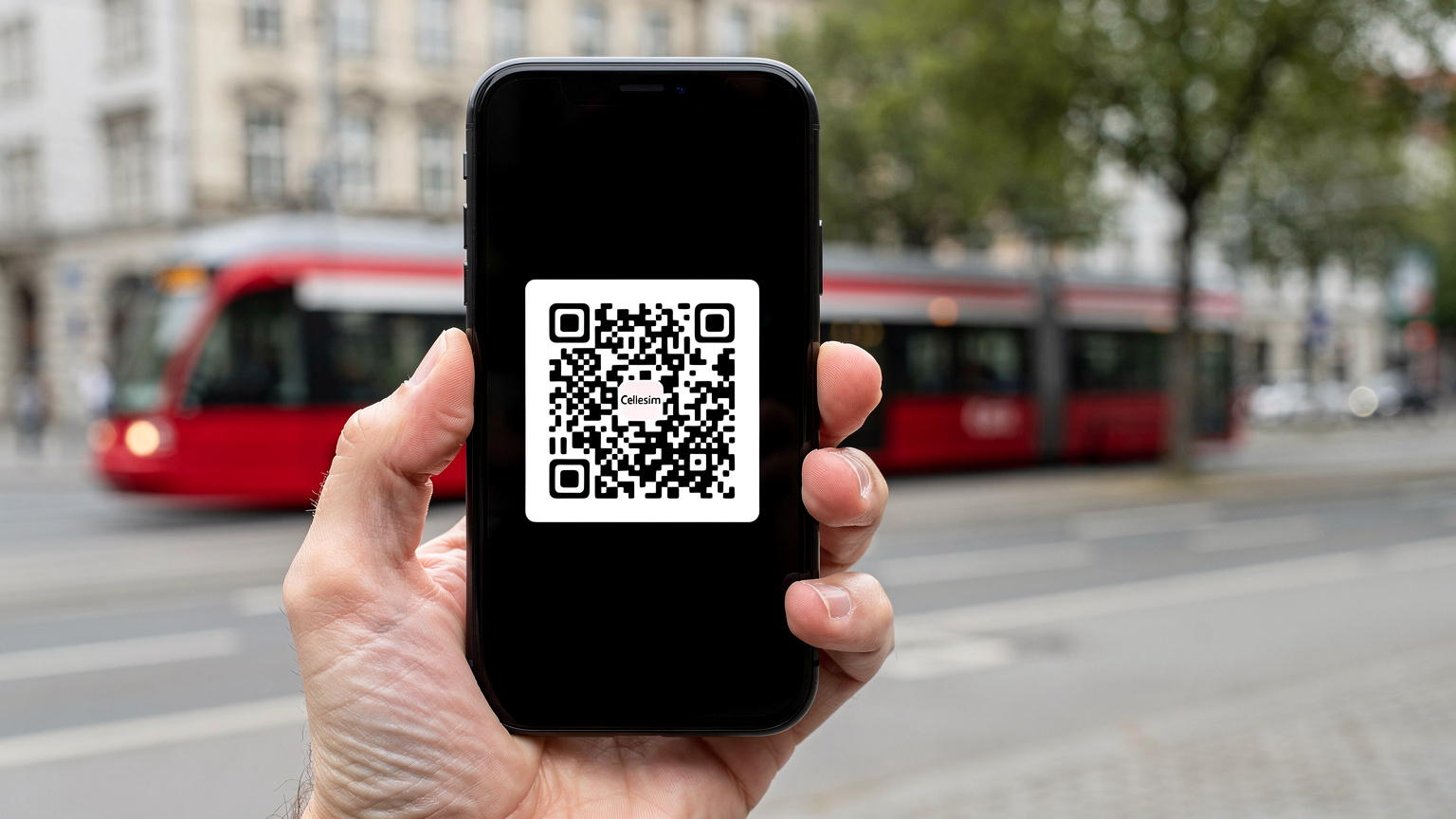 O mână umană ținând un smartphone, afișând un cod QR Cellesim pentru instalarea eSIM, în fundal, estompat, se vede un peisaj urban modern din Germania, cu un tramvai trecând.