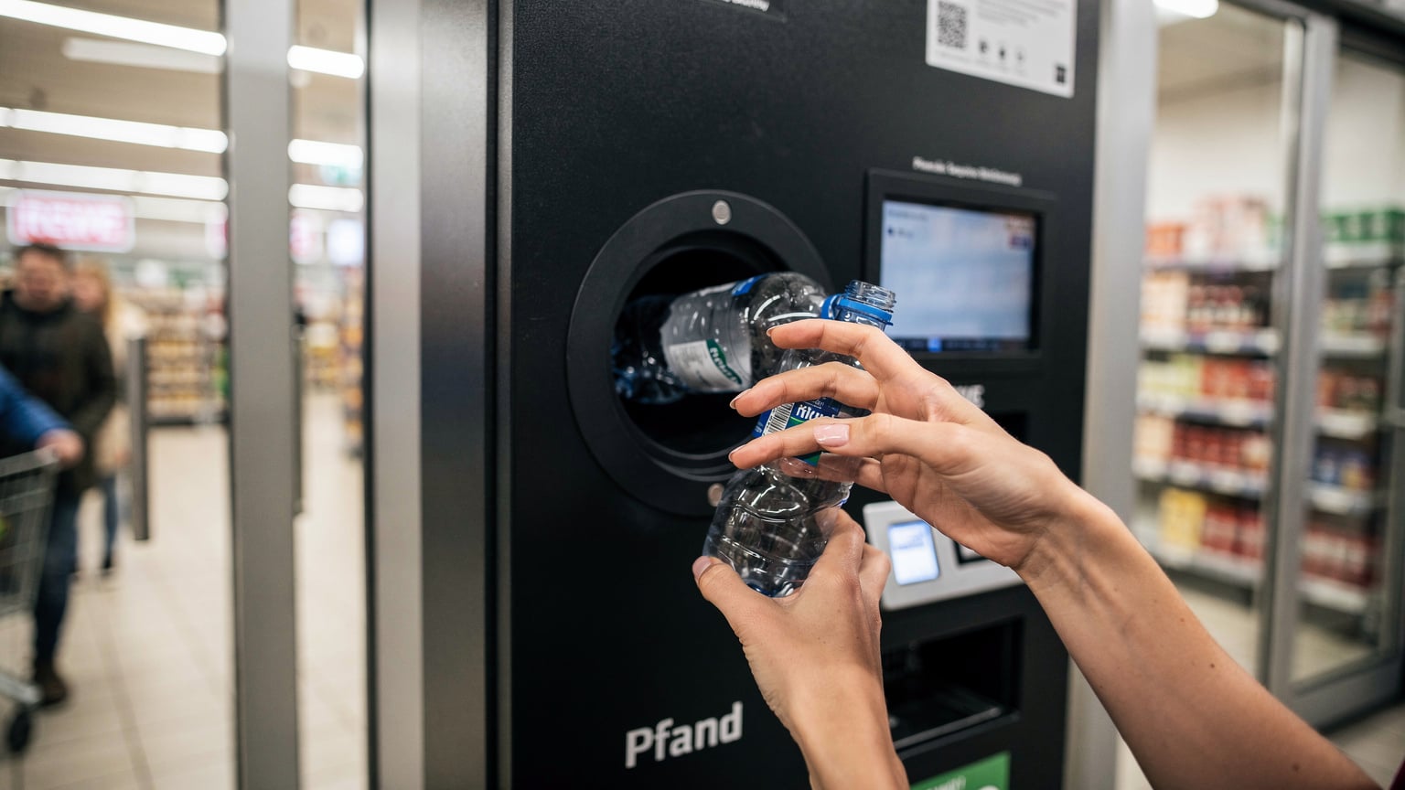 Une voyageuse dépose une bouteille en plastique vide dans une machine de recyclage Pfand à l'entrée d'un supermarché Rewe à Munich, un geste écologique devenu routine.
