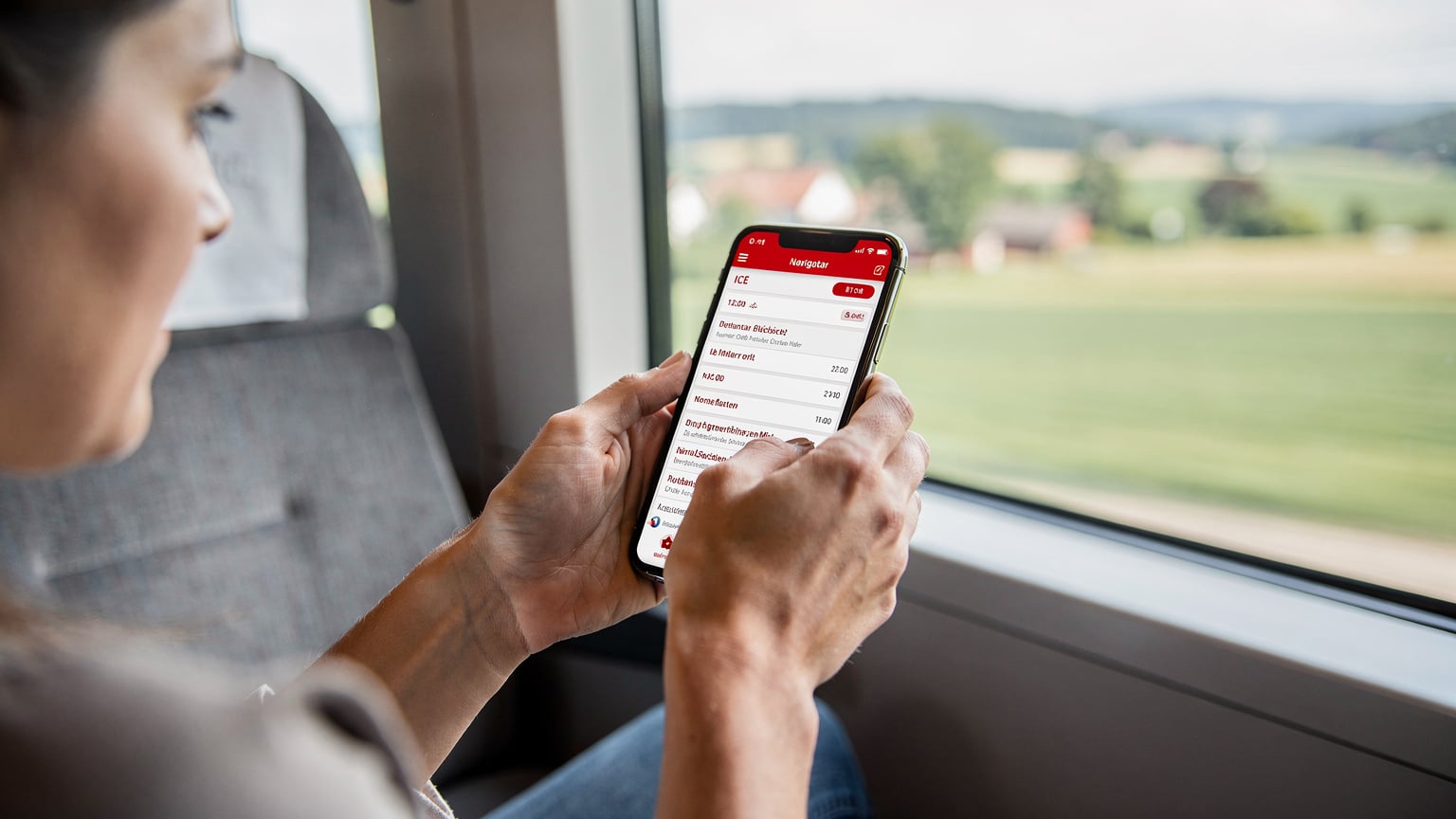 Gros plan sur un smartphone avec l'application DB Navigator affichant les horaires de train, tenu par des mains féminines dans un train ICE en mouvement, avec la campagne allemande floue en arrière-plan.