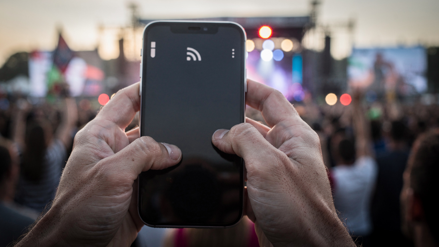 Zblízka na ruce držící telefon, kde se na obrazovce zobrazuje ikona slabého signálu na pozadí pulzujícího davu na festivalu.