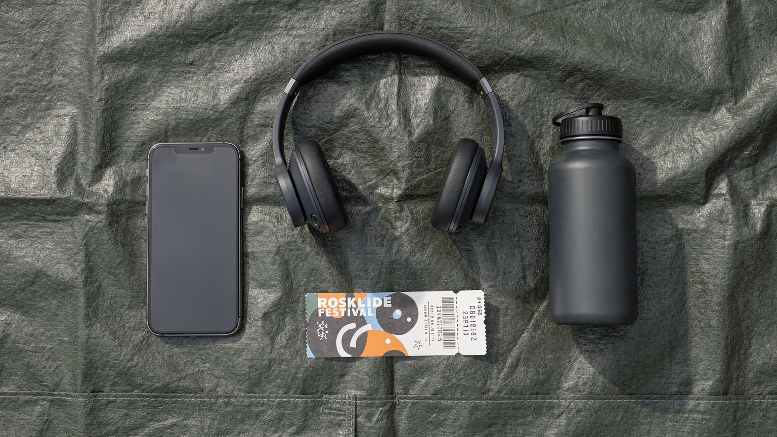 Et editorial flat-lay med en smartphone, et par over-ear hovedtelefoner, en festivalbillet, og en lille, genanvendelig vandflaske, alt sammen arrangeret på en robust, vandtæt dug.