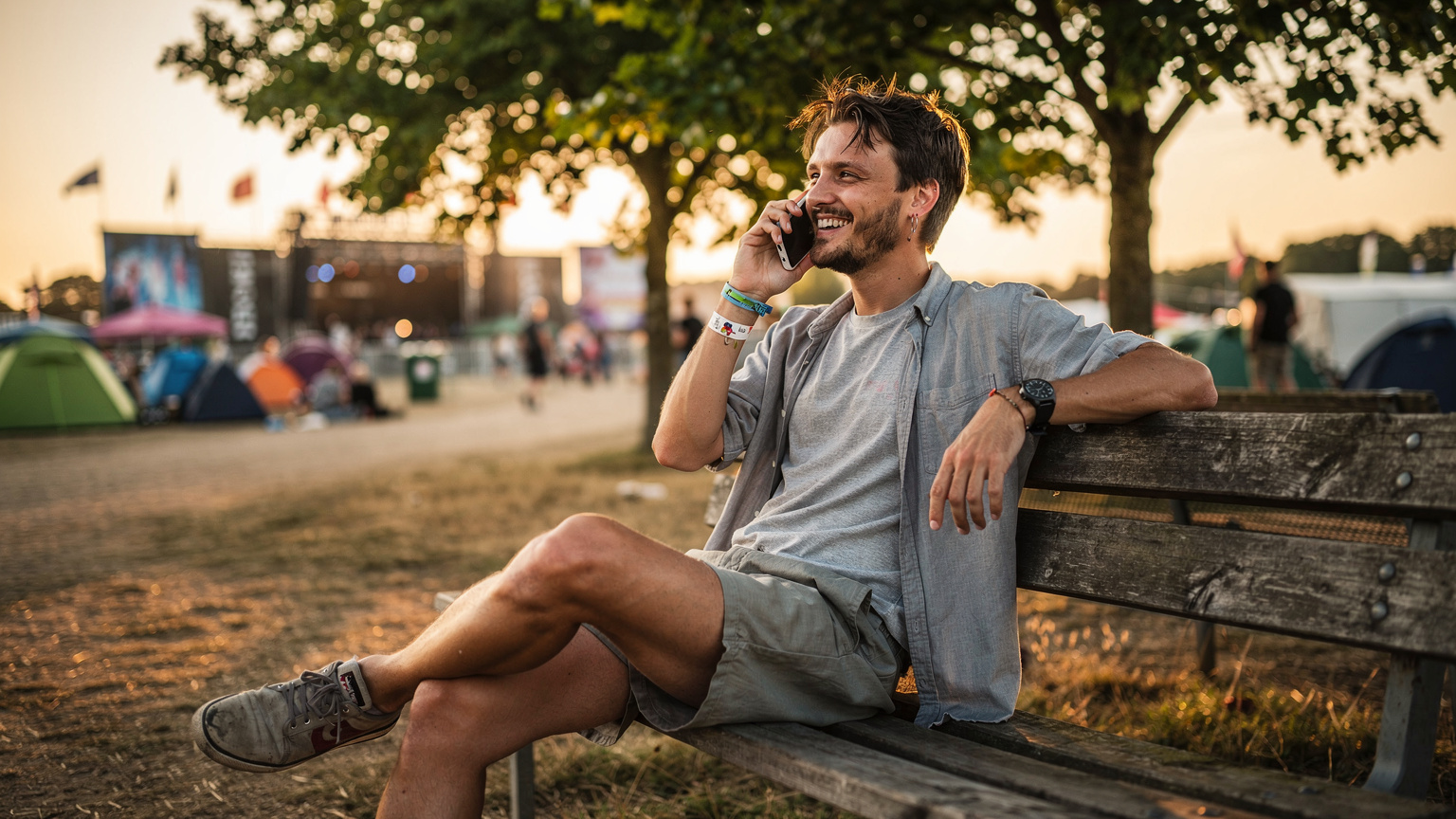 En ung mand i festivaltøj sidder på en træbænk og smiler, mens han taler i telefon, i baggrunden er der en rolig pause mellem koncerterne.