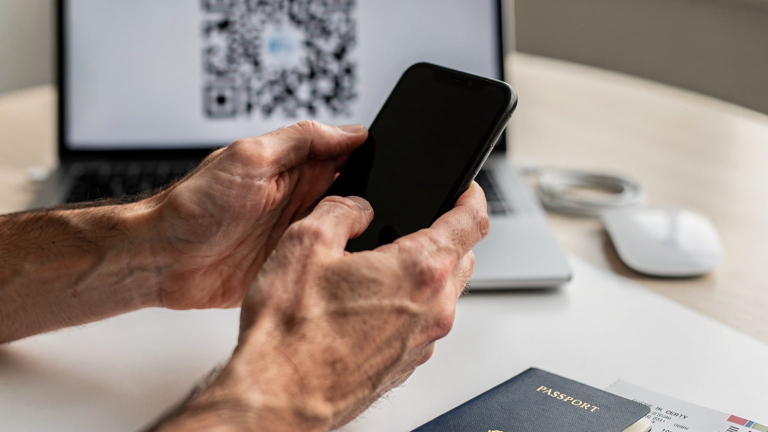 Close-up of hands holding a smartphone, with a QR code displayed on a computer screen in the background, showing eSIM activation.