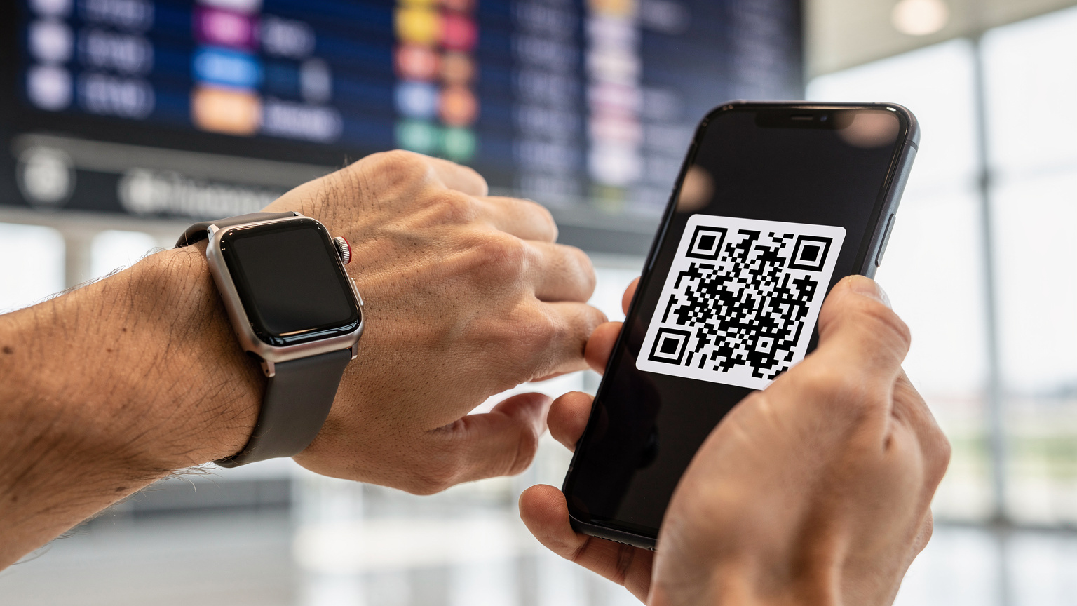 Zblízka záběr rukou člověka, který na letišti v Praze Ruzyni drží Apple Watch a skenuje QR kód eSIM ze svého telefonu. V pozadí je vidět odletová tabule.