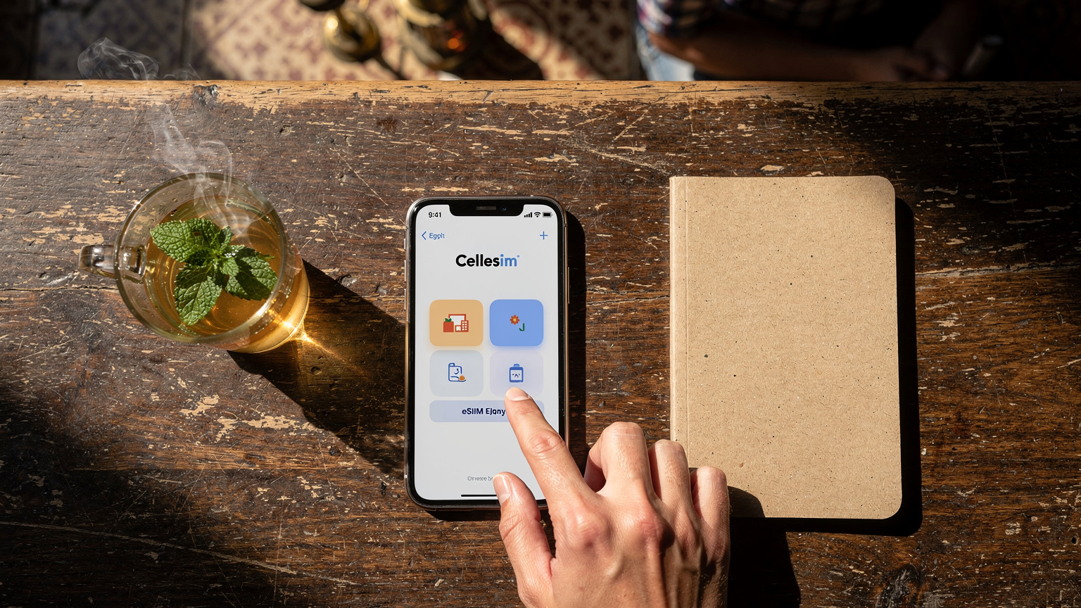 Một bàn tay đang nhẹ nhàng chạm vào màn hình điện thoại iPhone, hiển thị một ứng dụng Cellesim với các gói eSIM cho du lịch Ai Cập, đặt trên một chiếc bàn gỗ với một cốc trà bạc hà và một quyển sổ nhỏ, trong một không gian yên tĩnh và đầy nắng ở Cairo, Ai Cập.