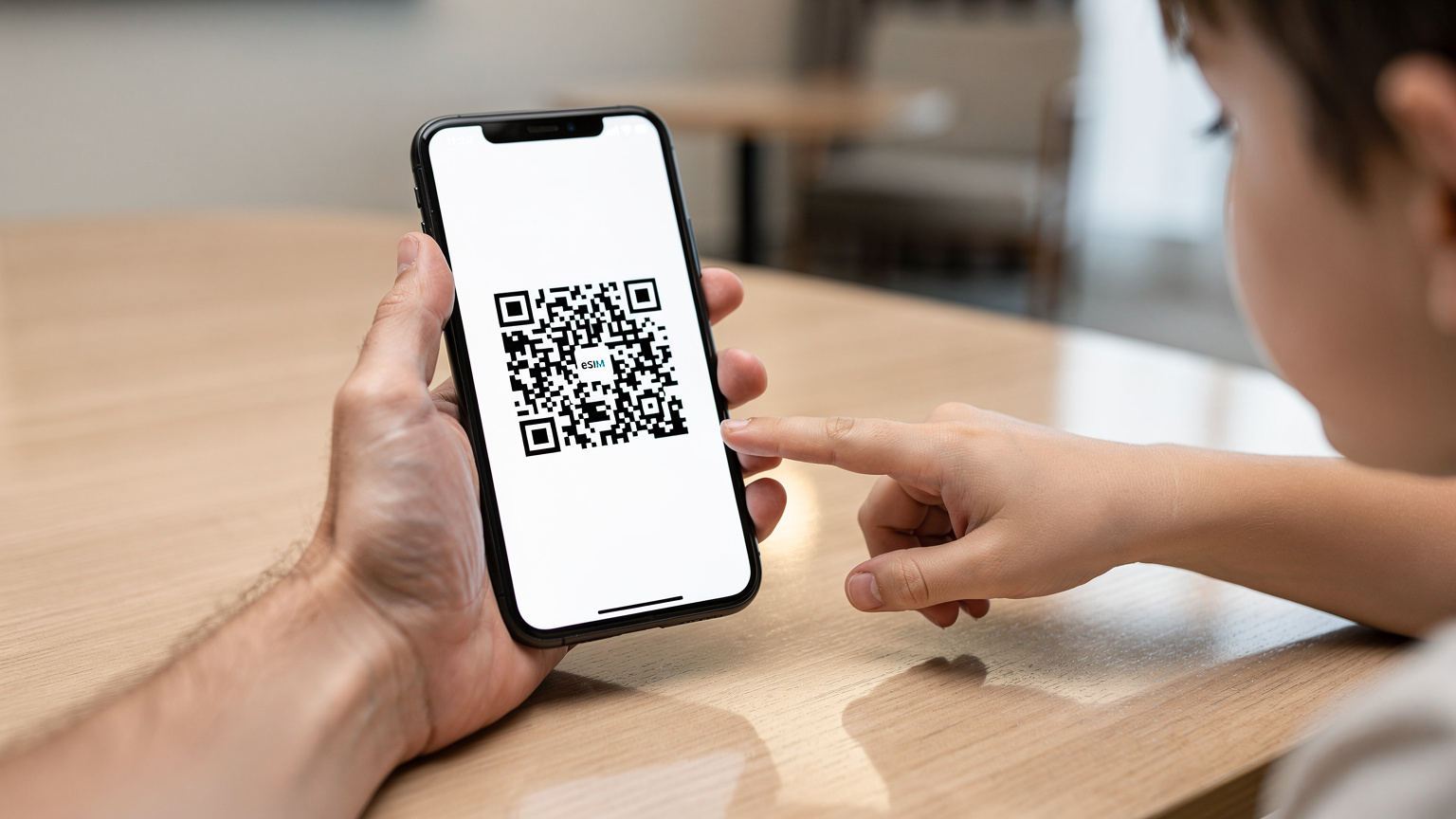Una mano adulta tiene uno smartphone con un codice QR per la scansione eSIM, mentre una mano di bambino indica lo schermo, su un tavolo di legno in un hotel moderno.