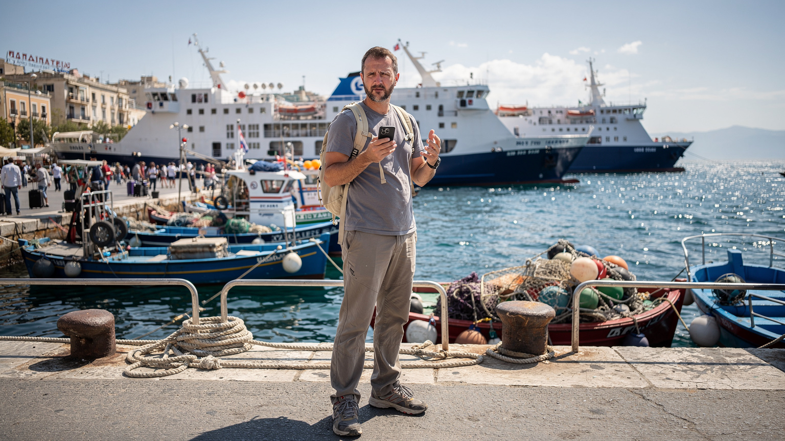 Un homme d'âge moyen, avec un sac à dos léger, fronce les sourcils en regardant son smartphone, cherchant une connexion Wi-Fi ou un signal, dans un port grec animé avec des ferries et des bateaux de pêche en arrière-plan.