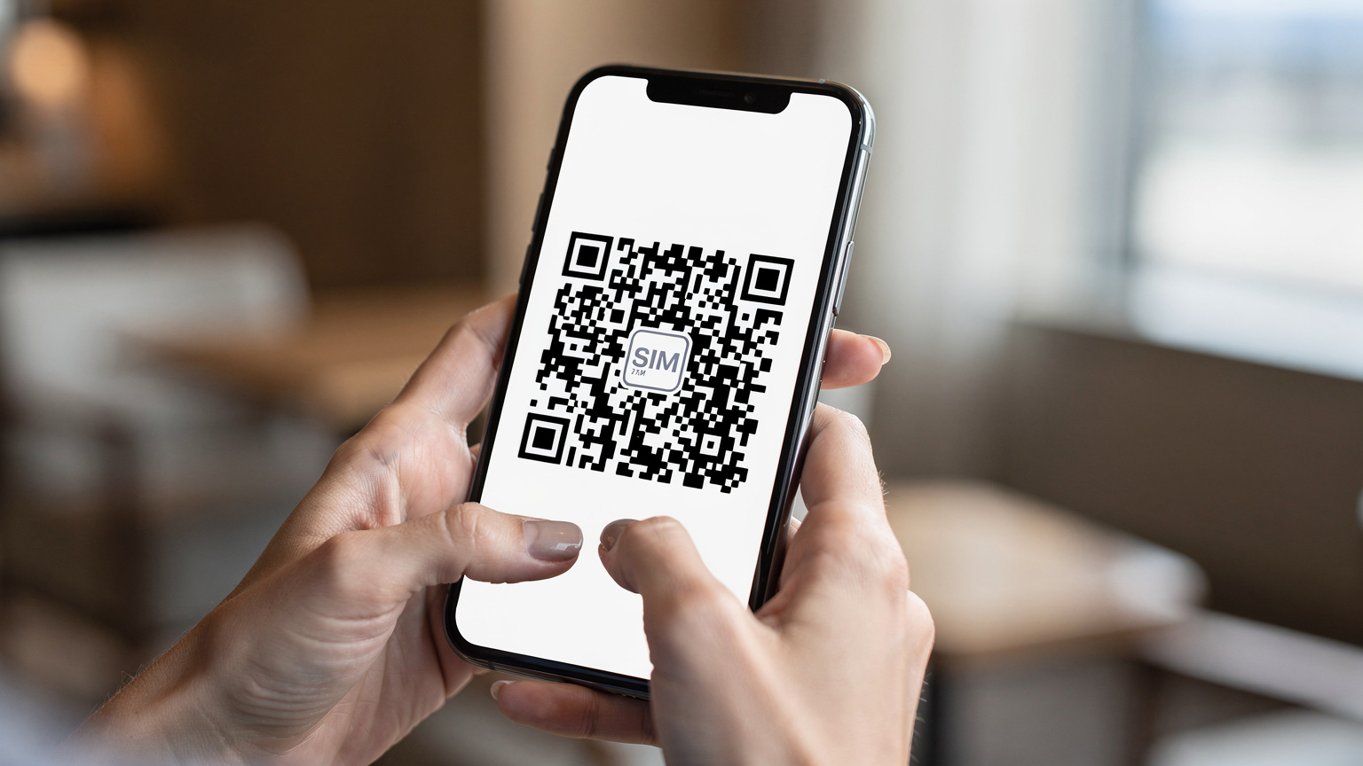 Un primo piano delle mani di una donna che sta digitando su un moderno smartphone, con un codice QR di un'eSIM visibile sullo schermo. Lo sfondo è sfocato e suggerisce un ambiente di viaggio, come la hall di un hotel o un caffè internazionale.