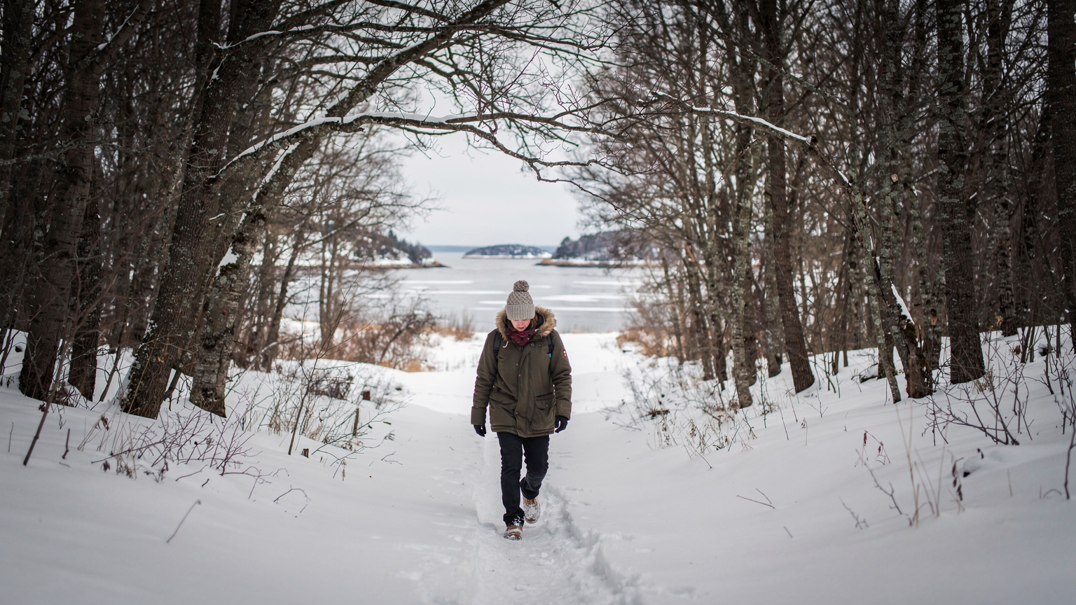 A lone traveler bundled in winter clothing carefully navigating a snow-covered, winding path through a sparse forest in the Aland Islands.