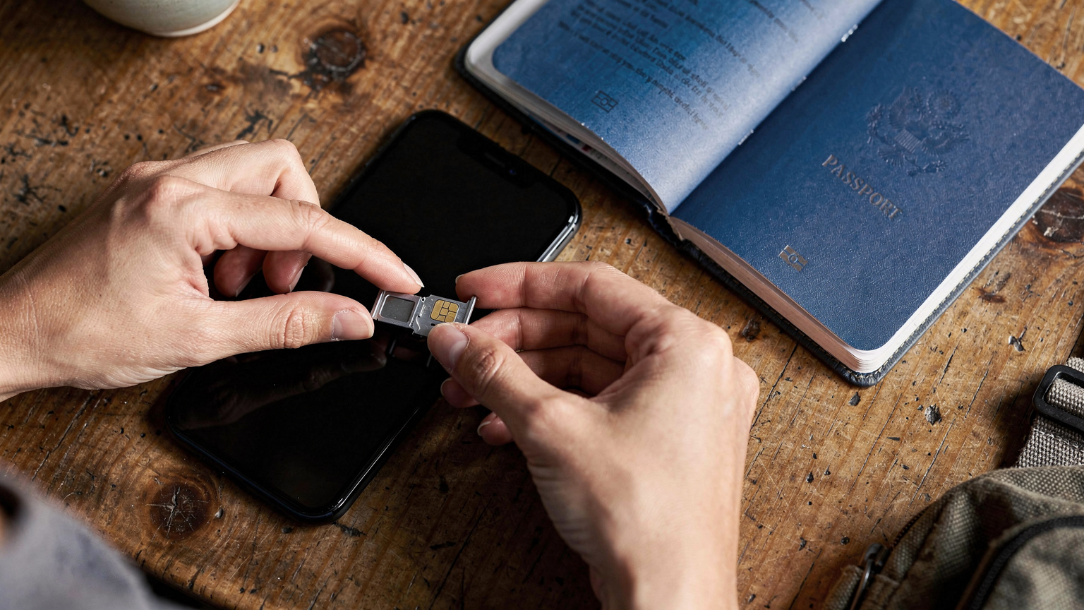 Close-up overhead shot of a traveler's hands gently placing a physical SIM card into a phone tray next to a passport, showing the detailed process of traditional SIM installation.