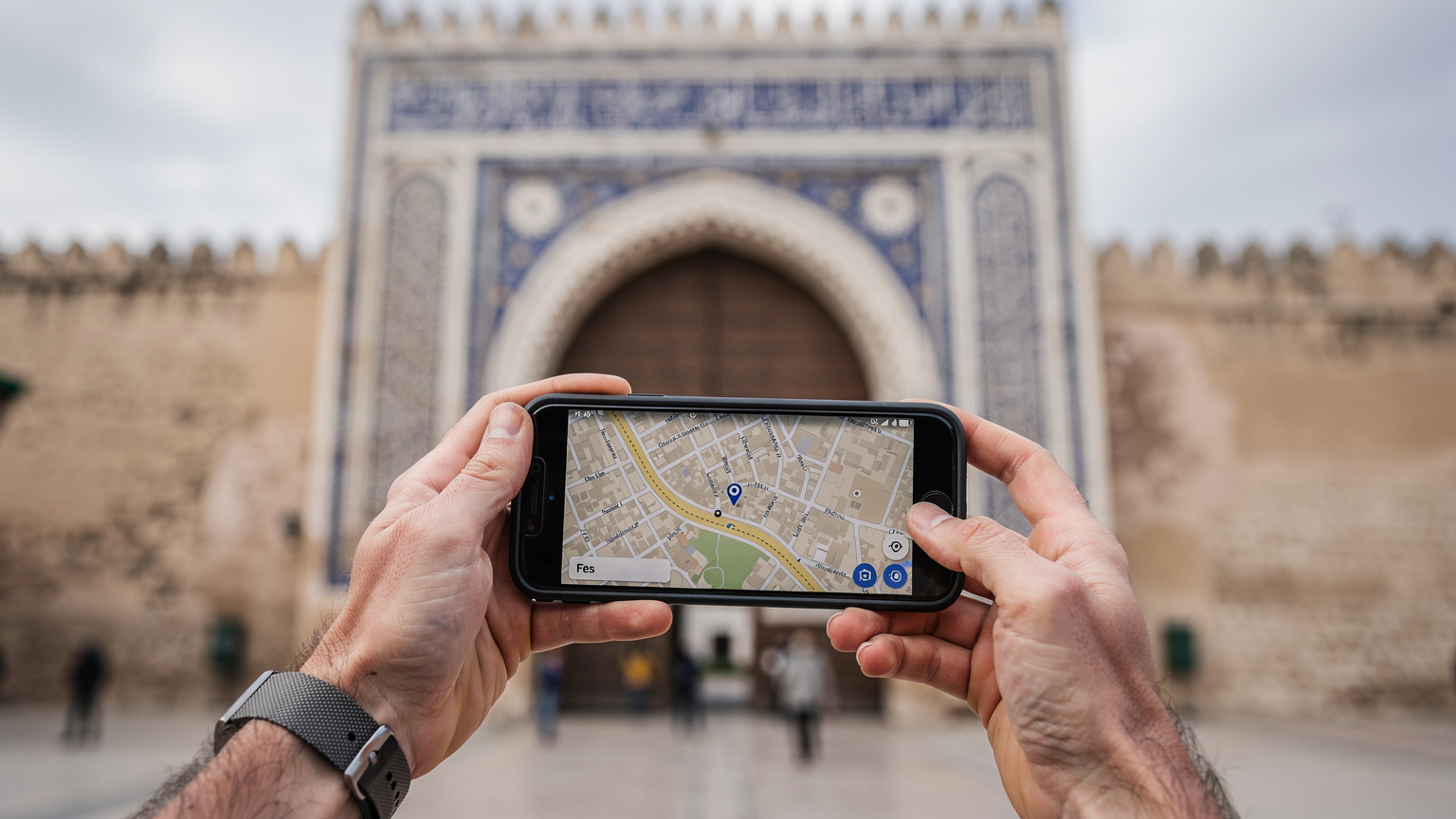 Ręce turysty trzymające smartfon, na ekranie którego widać mapę Fezu z aktywną nawigacją, w tle historyczna brama medyny.