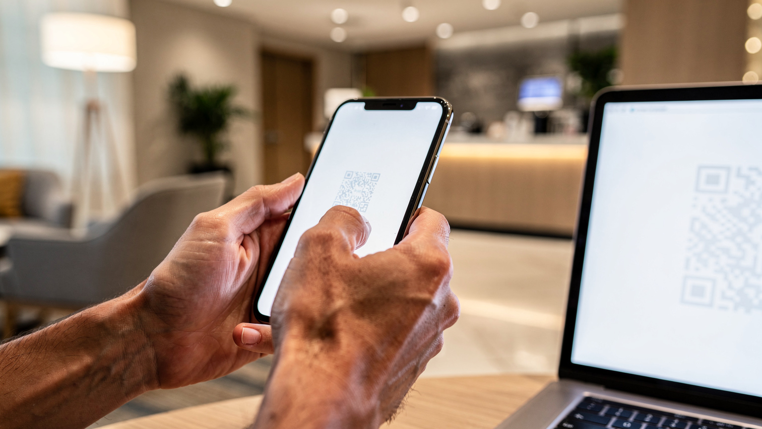 Närbild på händerna på en person som håller upp en smartphone för att skanna en QR-kod från en annan skärm, i en hotellobby.