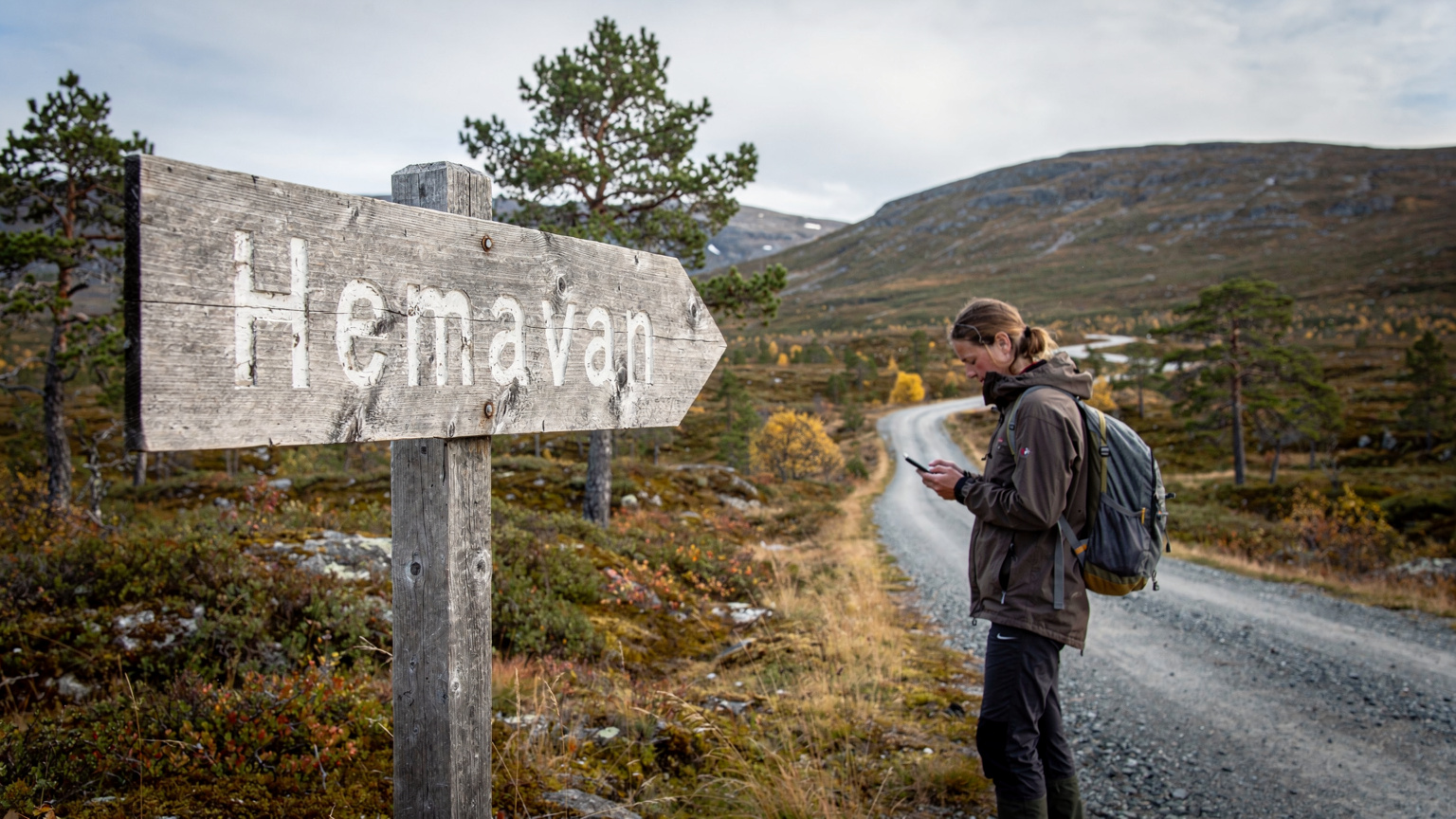 En vägskylt till en liten, pittoresk by i de svenska fjällen, med en person i bakgrunden som tittar på sin smartphone.