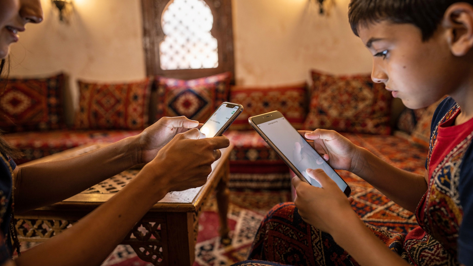 Une mère montre à son enfant comment activer un point d'accès personnel sur son smartphone pour connecter la tablette de l'enfant, dans un salon marocain traditionnel avec des coussins colorés.