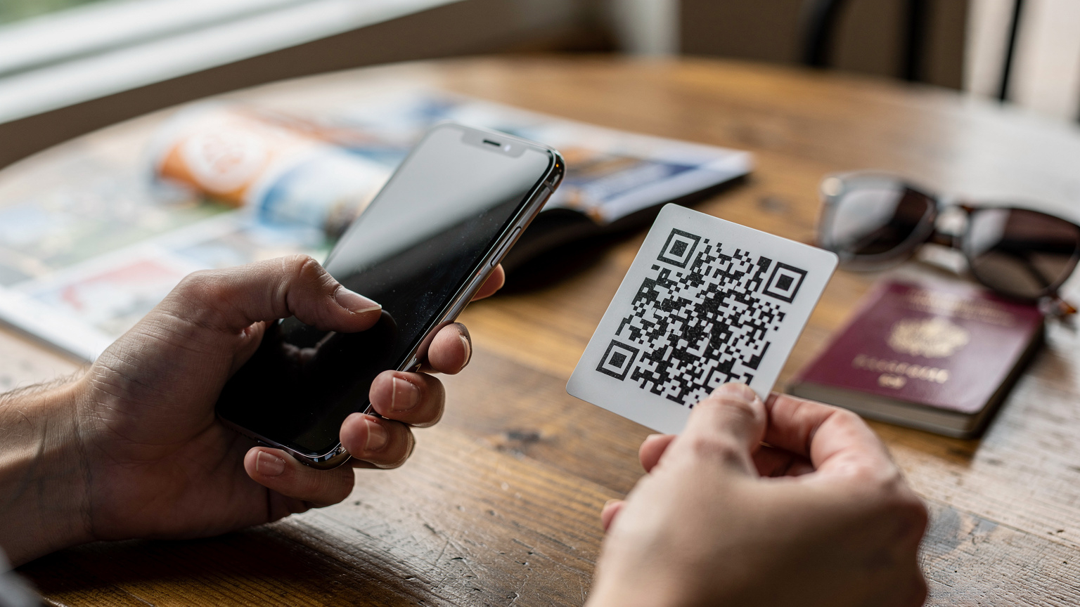 En nærbilde av hender som holder en smarttelefon og skanner en QR-kode for å aktivere et eSIM, med en reisebrosjyre og pass liggende i bakgrunnen på et kafébord.