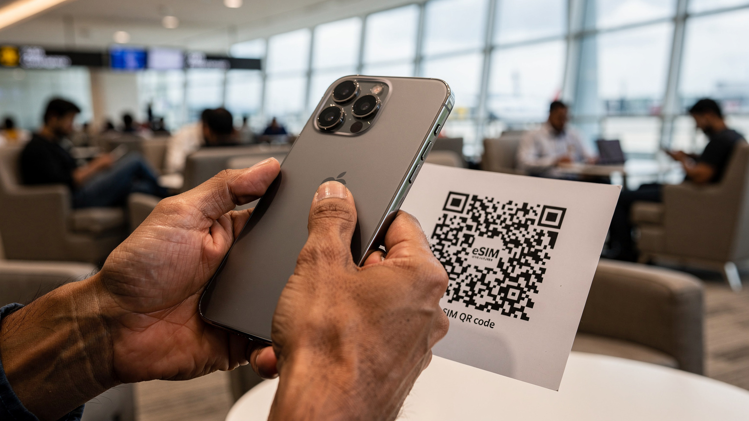 एक भारतीय यात्री अपने iPhone 18 पर eSIM QR कोड स्कैन कर रहा है, जिसके बैकग्राउंड में एक व्यस्त एयरपोर्ट लाउंज दिख रहा है, जहाँ अन्य यात्री अपने फोनों का उपयोग कर रहे हैं।