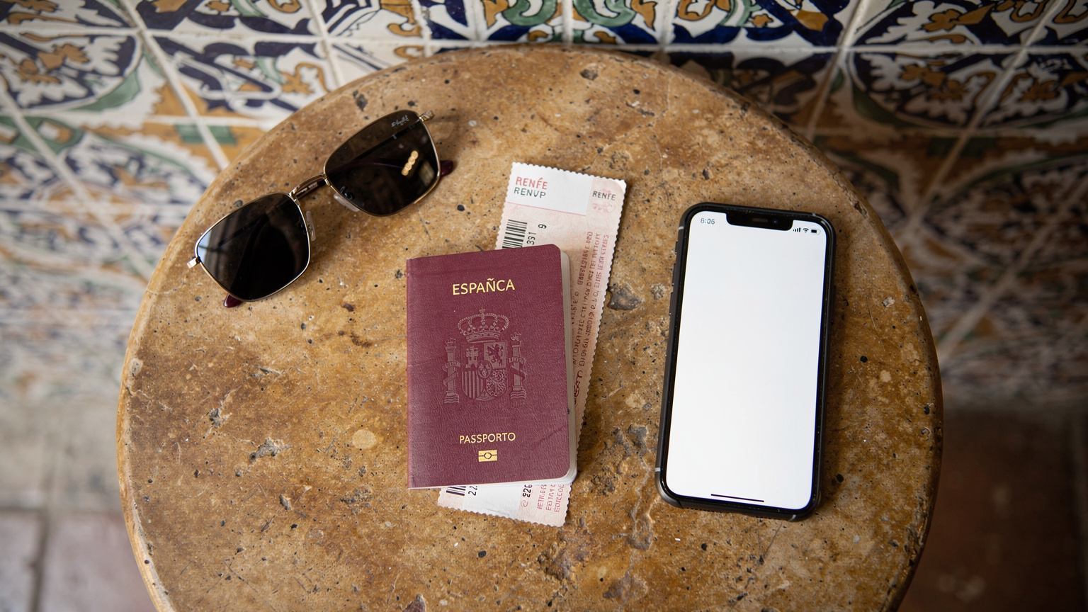 Un plano cenital de una mesa de café en la que se ven unas gafas de sol, un pasaporte español, un billete de tren de Renfe y un móvil con la pantalla encendida, con un fondo de azulejos andaluces.