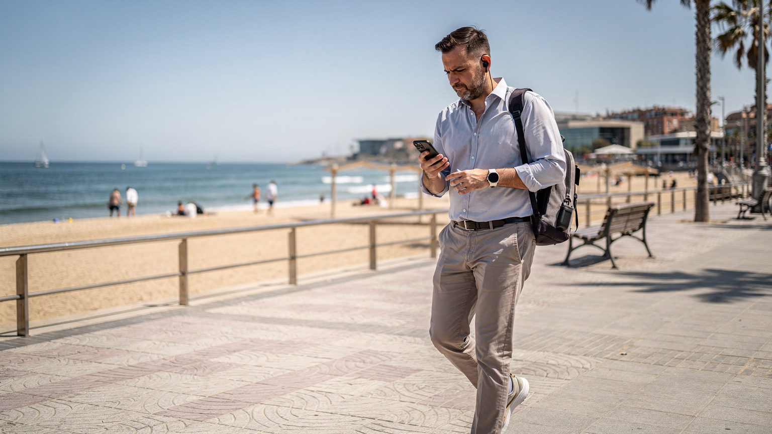 Un ingeniero de telecomunicaciones revisa los parámetros de su móvil mientras camina por el Paseo Marítimo de Barcelona, con la playa de la Barceloneta de fondo.