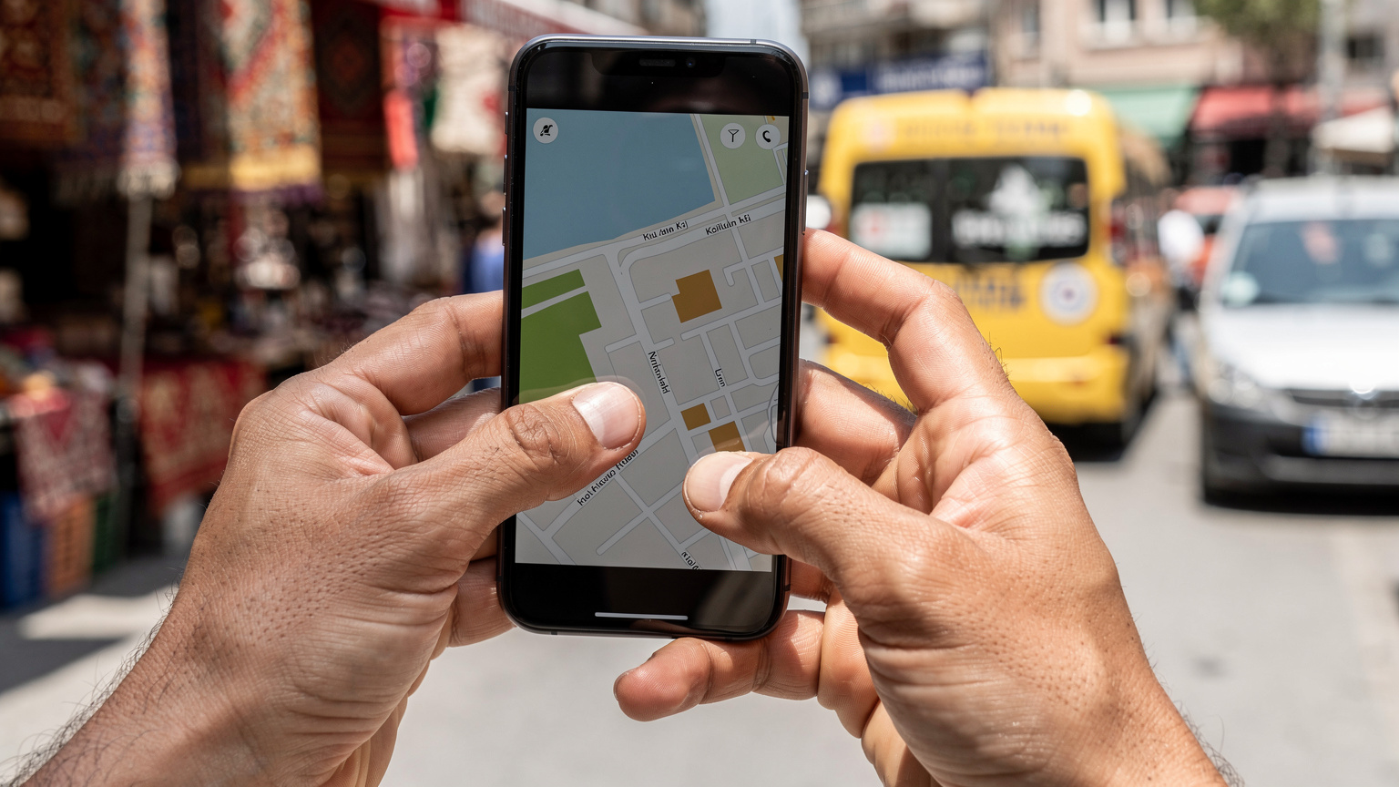 Nahaufnahme von Händen, die ein Smartphone halten, auf dem eine Karten-App geöffnet ist, während im Hintergrund ein belebter türkischer Markt oder eine Bushaltestelle zu sehen ist, was eine typische Reisesituation darstellt.