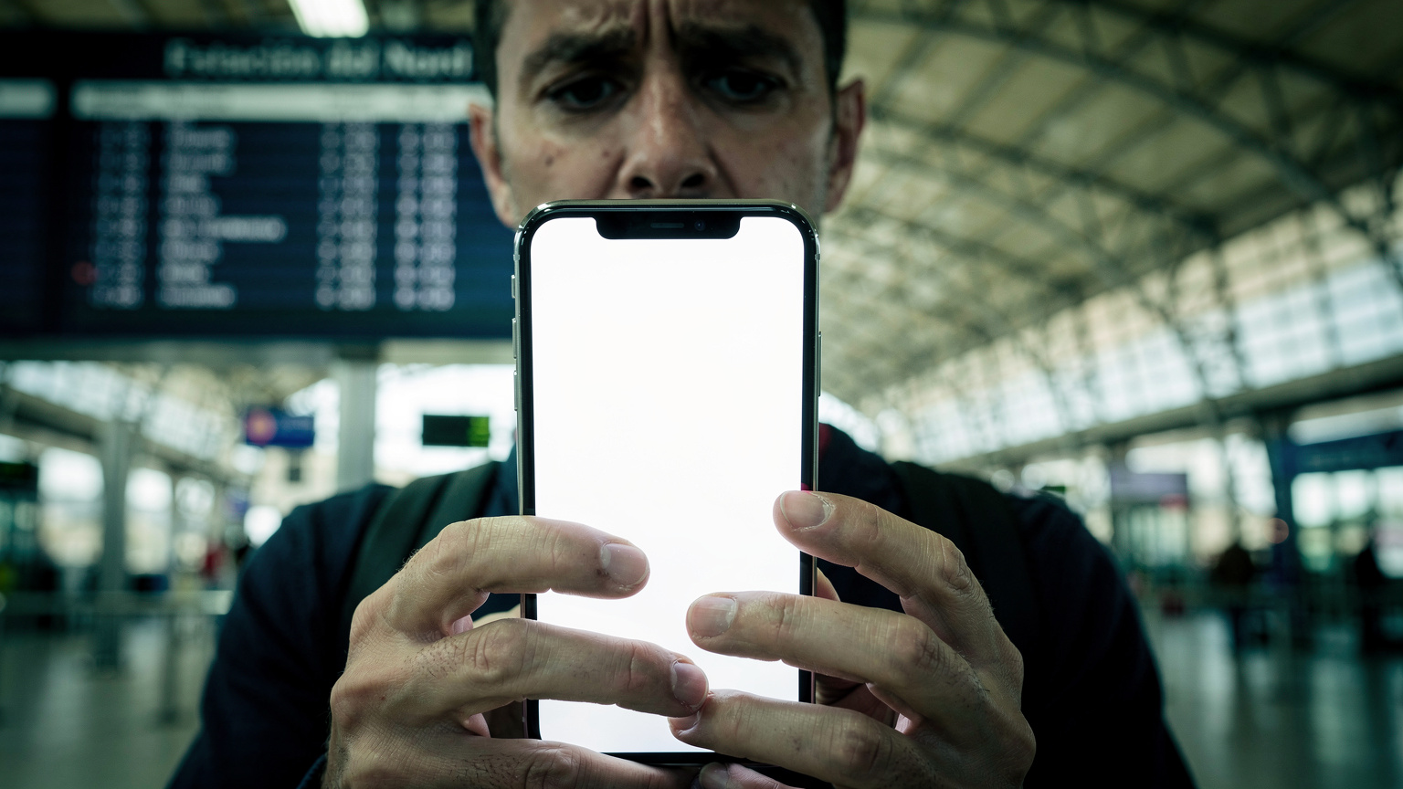 Un viaggiatore guarda preoccupato il suo smartphone, che mostra un avviso di dati in esaurimento, mentre si trova al terminal degli autobus di Barcellona, con un grande tabellone degli orari sullo sfondo. La scena è illuminata da luci fluorescenti.