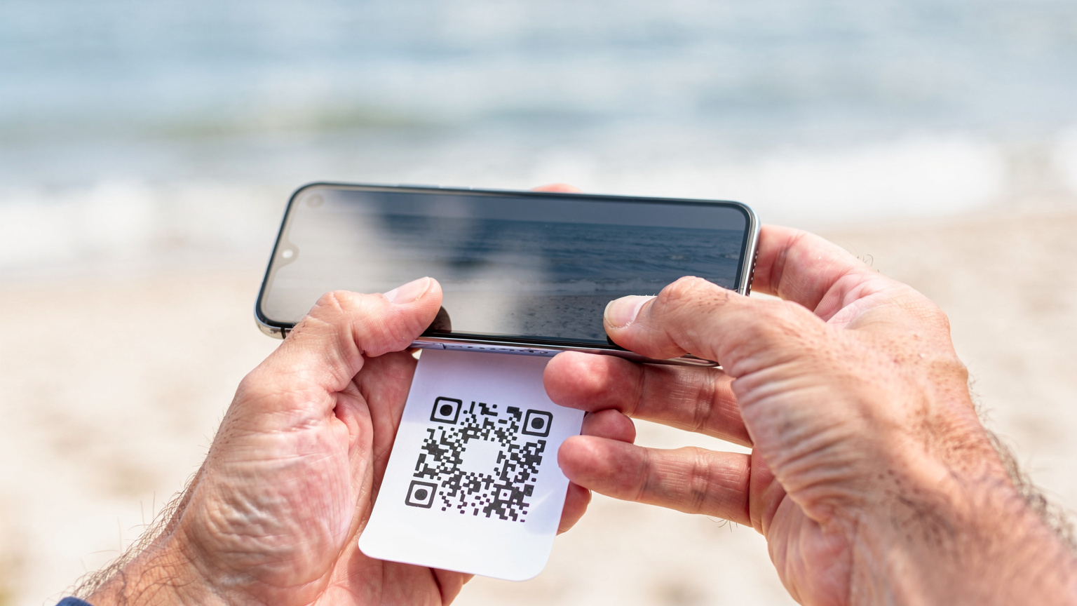Zbliżenie na dłonie rodzica skanującego kod QR eSIM na ekranie smartfona, w tle widać morze i plażę.