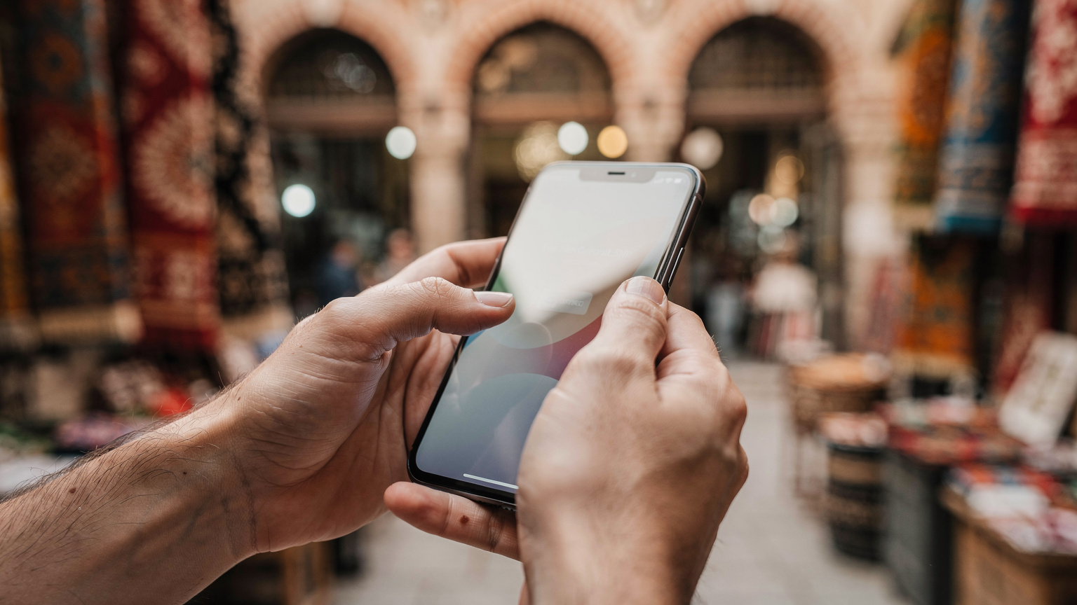 Bir seyyah, mobil bankacılık uygulamasından eSIM paketi satın alırken, parmaklarıyla akıllı telefon ekranında kaydırma hareketi yapıyor, arka planda bulanık bir şekilde tarihi bir çarşı görünüyor.