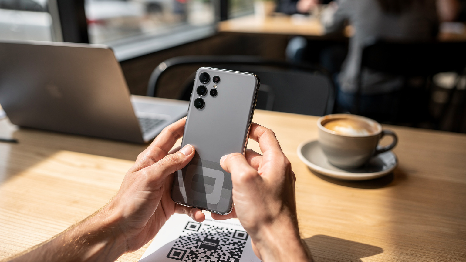 Manos de un viajero sosteniendo un Samsung Galaxy S26 en una cafetería moderna en EE. UU., mostrando el proceso de escaneo de un código QR para activar una eSIM. El fondo está ligeramente desenfocado, con tazas de café y un ordenador portátil.