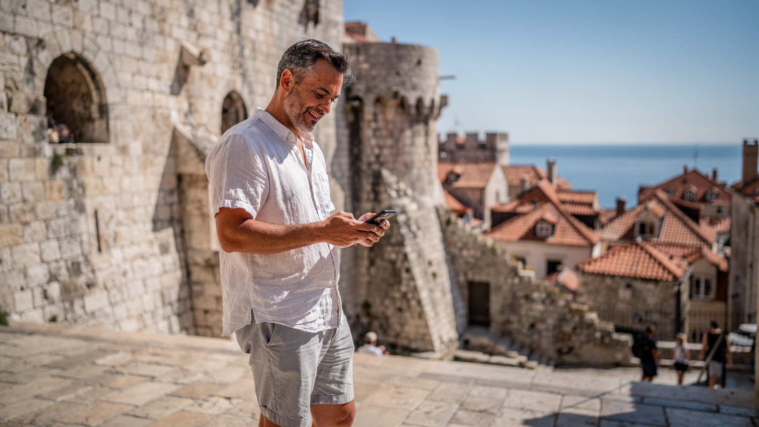 En mann i 40-årene, kledd i lette sommerklær, står foran den gamle bymuren i Dubrovnik, Kroatia, og ser på telefonen sin for å navigere. Solen skinner på de historiske bygningene.