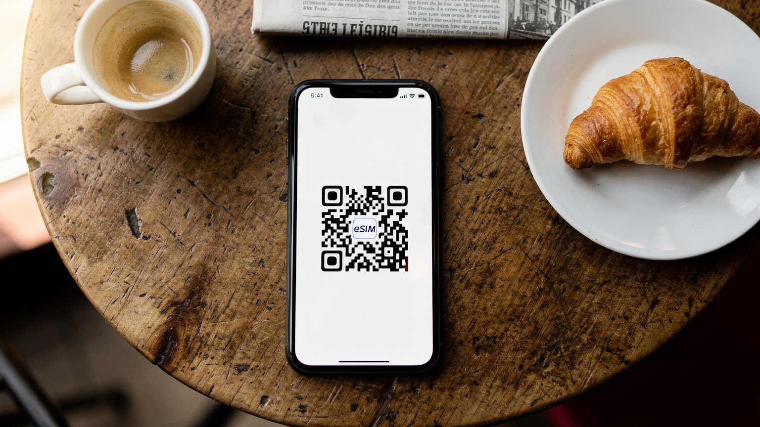 Un smartphone avec un QR code eSIM affiché, posé sur une table de café parisien en bois, avec une tasse de café et une petite viennoiserie en arrière-plan.
