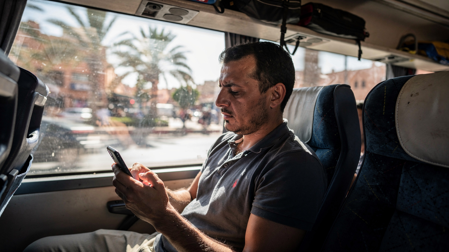 Zamyślony podróżnik wpatrujący się w ekran smartfona w zatłoczonym autobusie CTM w Maroku, z widocznym na zewnątrz dynamicznym krajobrazem miejskim.