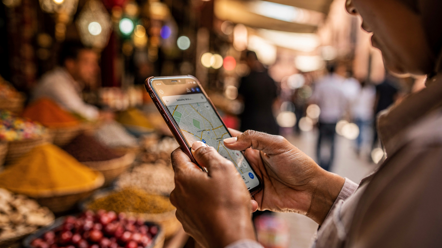Tangan seorang ibu dengan cekatan menyentuh layar smartphone-nya yang menampilkan aplikasi peta offline, di tengah keramaian pasar tradisional di Marrakech, Maroko, menunjukkan persiapan matang seorang traveler keluarga.