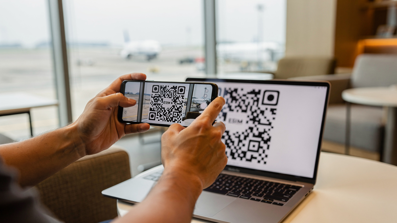 Một du khách đang scan mã QR eSIM trên màn hình laptop bằng điện thoại của mình trong sảnh chờ sân bay Tân Sơn Nhất, ánh sáng từ cửa sổ lớn chiếu vào.