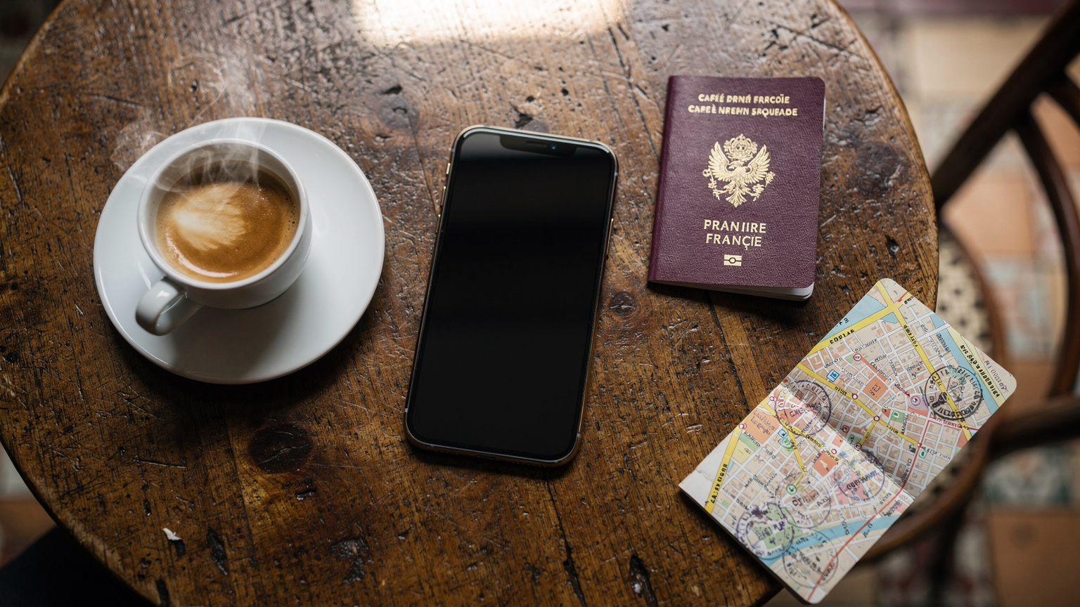 Une vue aérienne d'un iPhone posé sur une table de café en bois, à côté d'une tasse de café fumante et d'un passeport français ouvert, symbolisant la connectivité essentielle pour le voyageur moderne.