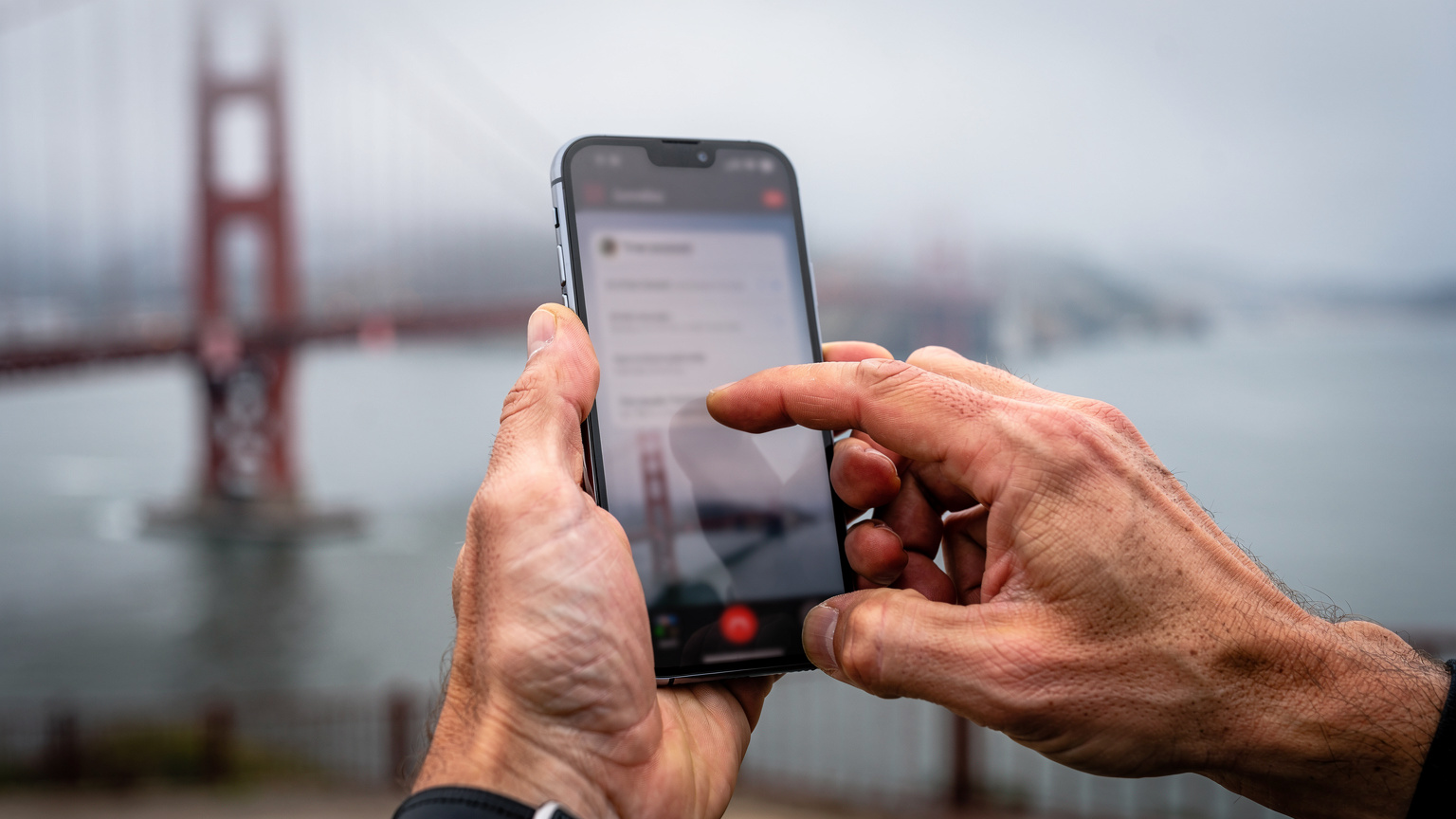 Detailní záběr rukou cestovatele, který na svém smartphonu ověřuje pokrytí sítě, zatímco stojí před ikonickým mostem Golden Gate v San Franciscu.