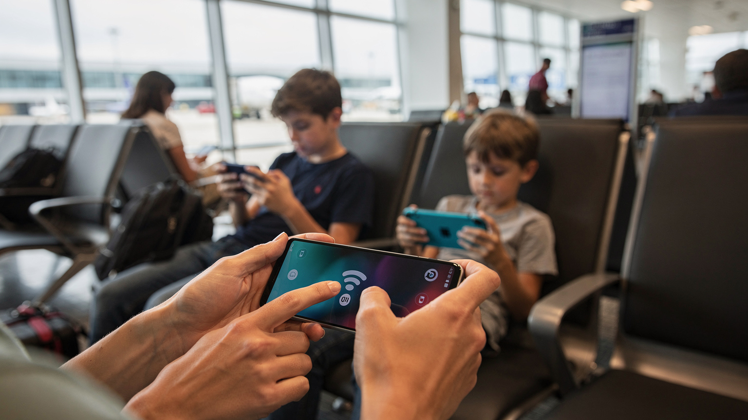 Mãos de uma mãe apontando para o ícone de Wi-Fi em um smartphone, enquanto um adolescente e uma criança pequena jogam em seus próprios dispositivos em uma área de espera de aeroporto.
