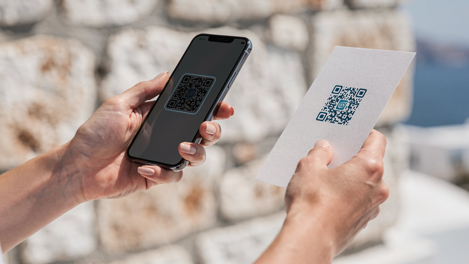Κοντινό πλάνο ενός χεριού που σαρώσει έναν QR κωδικό eSIM σε ένα smartphone, με φόντο έναν παραδοσιακό τοίχο από πέτρα σε ένα ελληνικό νησί.