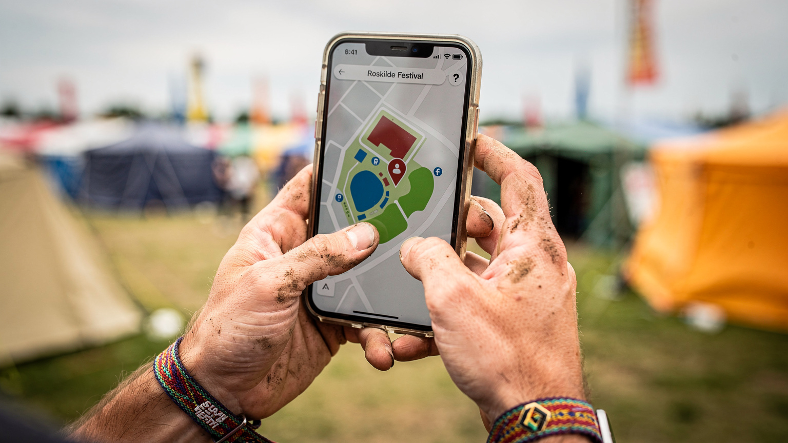 En tæt in-situ optagelse af en festivaldeltagers hænder, der holder en smartphone med et kort over festivalpladsen, der viser GPS-tracking af en ven.