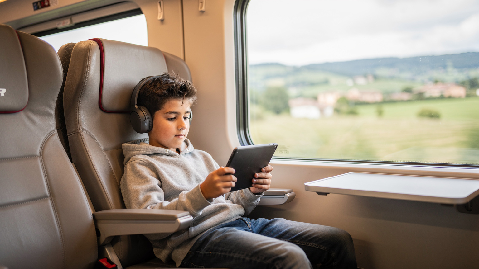 Un bambino di circa 8 anni, seduto in un moderno treno Frecciarossa, è completamente assorto in un gioco educativo sul suo tablet, indossando cuffie wireless. Fuori dal finestrino, il paesaggio italiano sfuma velocemente, suggerendo un viaggio.