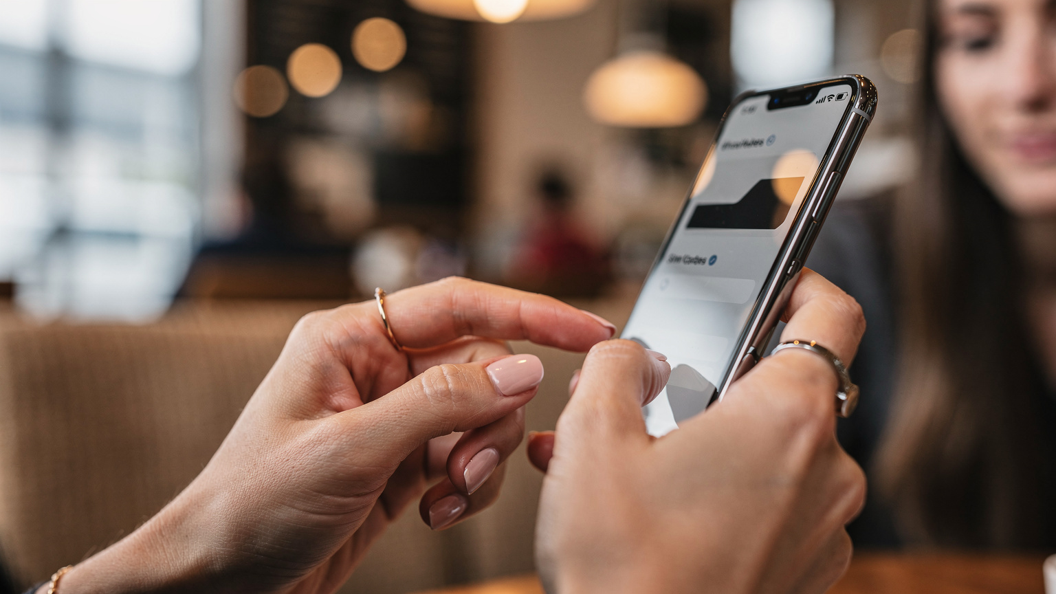 Gros plan sur les mains d'une femme utilisant un smartphone, l'écran affichant des réglages de carte SIM avec des options d'activation et de gestion de l'eSIM. L'arrière-plan, flou, suggère un environnement de café ou de lounge d'aéroport.