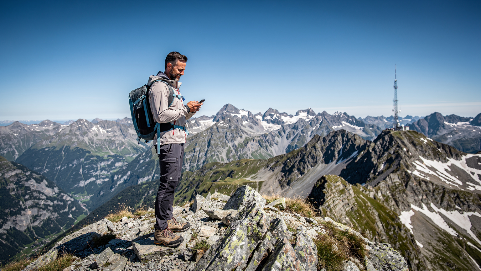 Un homme en tenue de randonnée, debout au sommet d'un col des Alpes françaises, consultant son smartphone avec une vue panoramique sur les sommets. Une petite antenne-relais de télécommunications est visible discrètement à l'arrière-plan.